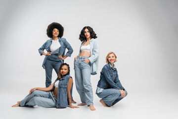 fashion shoot of multiracial girlfriends in trendy denim clothes posing barefoot on grey backdrop