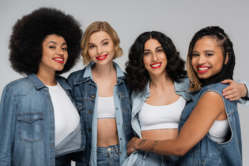 group of smiley multicultural girlfriends in denim clothes looking at camera on grey, diversity