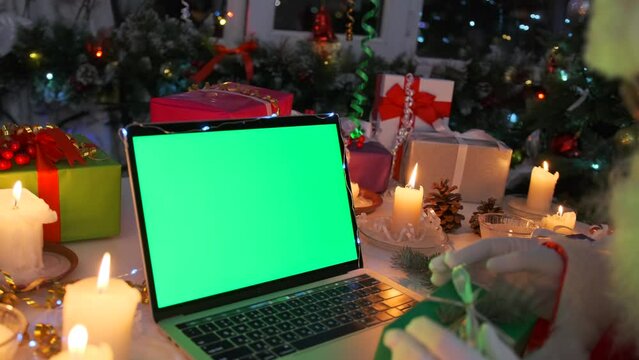 Grandfather Santa Claus sitting at laptop with mock up chroma key display screen, celebration gift in his hands next to candles, tinsel and flickering garlands of a Christmas tree. 