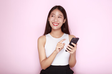 Fototapeta premium Standing and pointing happily, Portrait of a beautiful young woman in a light pink background, Asian woman holding a phone in one hand.