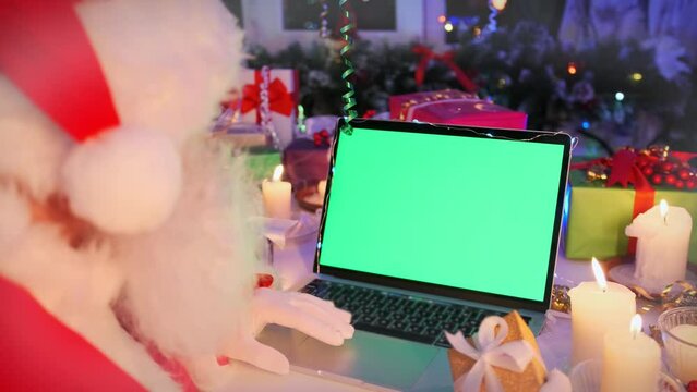 Bearded Santa Claus using laptop computer with green chroma key screen, burning candles, present boxes, tinsel and Christmas tree. Holidays and celebrations time.