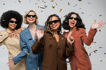 party time, cheerful multiracial girlfriends in sunglasses and suits under festive confetti on grey