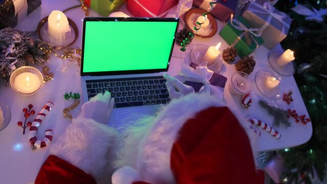 Grandfather Santa sits at laptop with chromakey on screen among Christmas decor of burning candles, packed gift boxes, fir branches and flickering garlands, close up, top view. Xmas spirit magic.