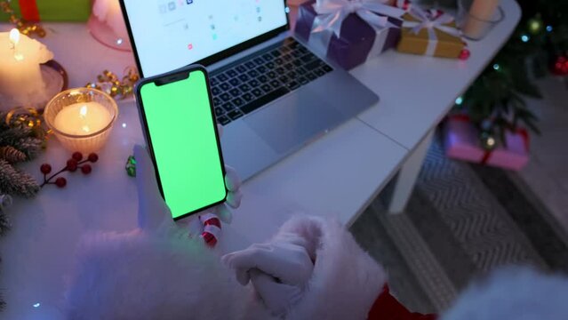 Smartphone with chromakey screen in Santa hands scrolls feed of news and social networks on background of desk with open laptop, lighted candles, gifts boxes, fir cones and branches in Christmas eve.