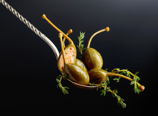 Pickled capers with thyme in spoon on a black background.