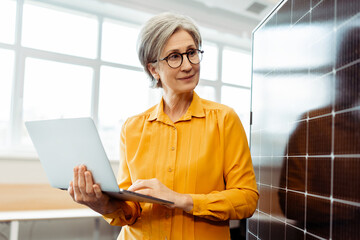 Successful smiling senior woman, engineer, developer holding laptop computer planning startup working with solar panel in office. Innovation, solar energy concept 