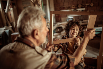 Fototapeta na wymiar Elderly carpenter making wood frame with little boy in workshop