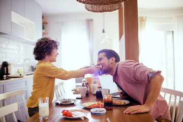Cute little boy playfully feeding father during breakfast in the kitchen