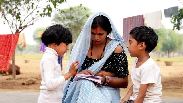 Indian village woman in saree teaching little daughter and son to read book, helping with primary school homework, showing pictures, telling fairy tale.