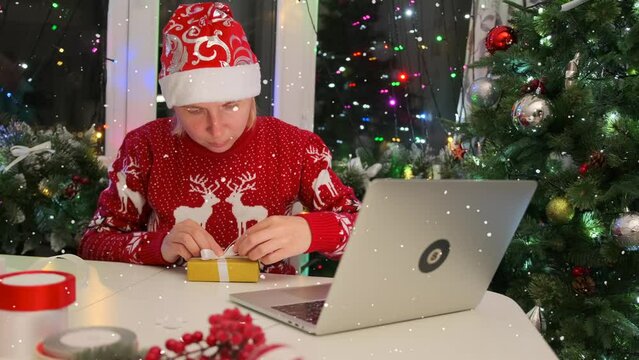 Christmas eve. Young woman in traditional red sweater and hat tying ribbon with bow on festive gift box, watch tutorial on laptop, against Xmas tree and decor. Wrapping New Year gifts. 