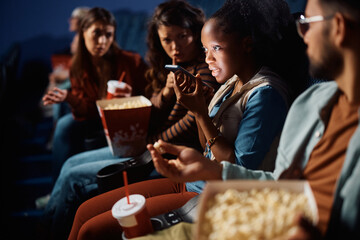 Black woman recording voice message on cell phone during movie projection in cinema.