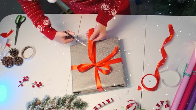 Female hands trim bow on wrapping  gift box, next to decor elements, wrapping paper, berries, pine cones, scissors, fir branches, Merry Christmas and Happy New Year 2024. Top view.