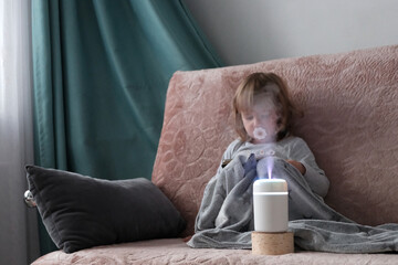 White aroma diffuser on the table. In the background is a blonde girl on the sofa, with a phone in...