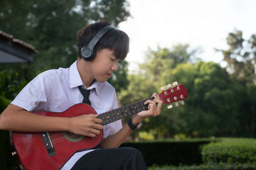 Asian cute boy holding acoustic guitar and ukulele and playing it under the tree in the park,...