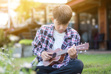 Asian cute boy plays ukulele in front yard of his house in the morning with the sunshine, soft and...