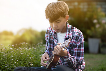 Asian cute boy plays ukulele in front yard of his house in the morning with the sunshine, soft and...