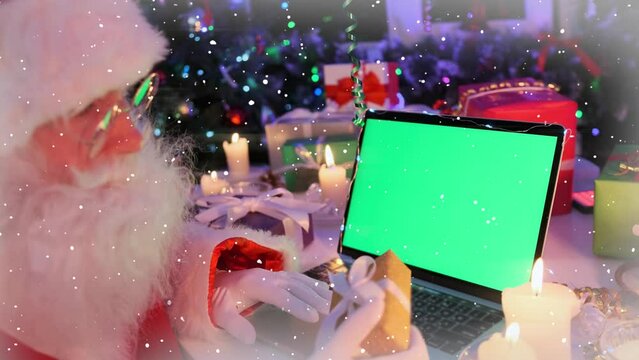 Funny Santa Klaus with beard and glasses sits at laptop with chromakey, congratulate children via video link, among gifts boxes, xmas tree, fir candles, cones, tinsel and garlands. Place promo sale ad