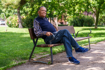 Relaxed senior man sits on a park bench, surrounded by trees, in casual attire and a contented...
