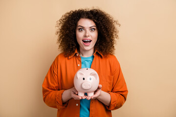 Fototapeta na wymiar Photo portrait of pretty young girl hold piggy bank excited impressed reaction wear trendy orange outfit isolated on beige color background