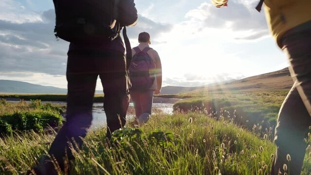 Three happy young hikers raise arms with joy and celebrate travel success standing near wild river at scenic mountain landscape. Motivation to tourist achievement