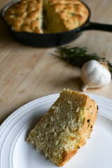 Slice of homemade focaccia bread on wood table with garlic, rosemary, and loaf in a cast iron pan in the background.