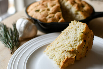 Slice of fresh baked, homemade focaccia bread on white plate with loaf in cast iron pan, garlic, and rosemary in the background