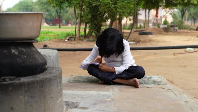 Little village girl doing homework in notebook and sitting on ground near goat farm. Indian countryside footage.