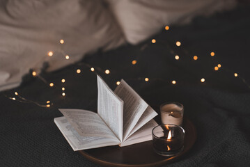 Open paper book and burning scented burning candle on tray in bed over Christmas lights in dark room close up. Winter holiday season.