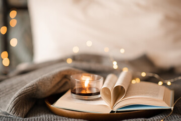 Open paper book with folded pages in heart shape and burning scented burning candle on tray in bed...