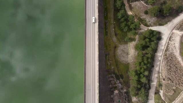 Static drone view from above of a road divides a reservoir and a wooded area through the center of the image and a vehicle drives from the bottom to the top.