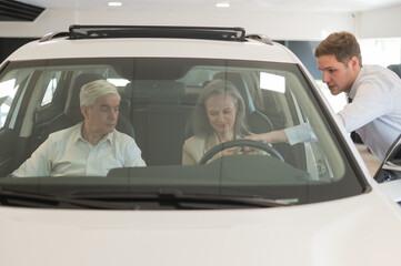 A gray-haired Caucasian woman is sitting behind the wheel of a car. Car dealership salesman consulting mature married couple.