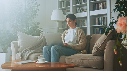 Obraz premium Young brunette woman uses a laptop notebook while sitting on the sofa in the living room at home. Businesswoman works remotely. Typing on a laptop keyboard while concentrating on work