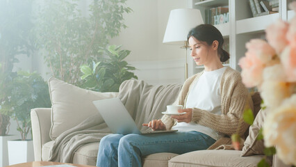 Portrait of Pretty Young Business Woman Drinking Coffee And Working Notebook From Home in Cozy Sunny Living Room. Young woman using laptop computer on sofa. Browsing on social media relaxed - Powered by Adobe