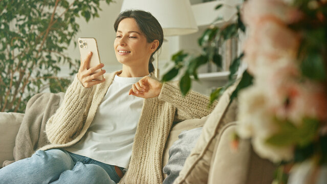 Young beautiful woman relaxing on the sofa, talking on the phone, smiling and contact. Mobile communication app using smartphone. Lady spending time at home with cell gadget technology
