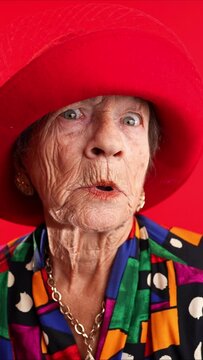 Vertical video of funny fisheye portrait of elderly senior old woman with wrinkled skin looks at camera gives great idea gesture showing explosion of thinking posing isolated on red background.