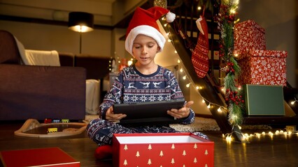 Little smiling boy in Santa's hat sitting on floor and playing video games on tablet computer in living room decorated for Christmas