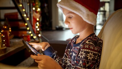 Closeup portrait of little boy in Santa's hat sitting on sofa and playing games on tablet computer...