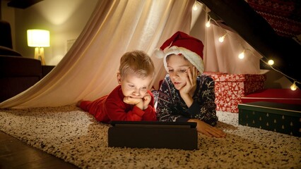 Two boys in pajamas lying under stairs at home and watching video on tablet computer while waiting for Christmas eve celebrations. Winter holidays, celebrations and party.