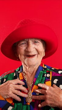 Vertical video closeup funny portrait of happy elderly toothless senior woman wearing red hat pointing fingers herself ask say who me, smiling talking and pointing. Isolated on red background.