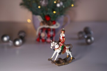 a toy figure of a nutcracker in a red suit on a white horse on the background of a vase with a bouquet of blue spruce branches with balls and glowing garlands on the table