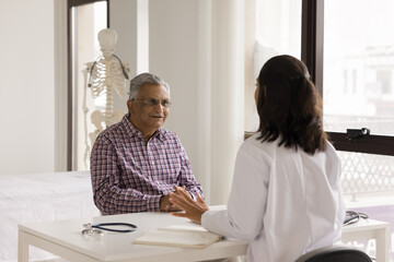 Positive senior Indian patient man consulting young doctor woman, talking to practitioner at hospital office workplace, discussing geriatric healthcare problems, illness, treatment