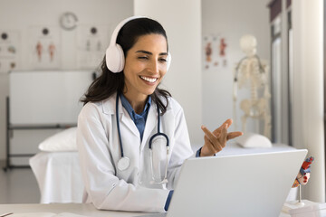 Obraz premium Happy beautiful young doctor woman in headphones talking to patient on video call, using laptop for online conference chat, giving professional consultation on Internet from hospital workplace