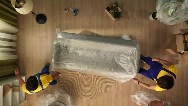Top view of new apartment living room. Delivery service male workers in uniform take out sofa wrapped in film.