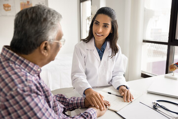 Positive empathetic young doctor woman giving support to elderly patient man with geriatric diseases, touching hand over workplace table, holding arm, expressing medical care, smiling - Powered by Adobe