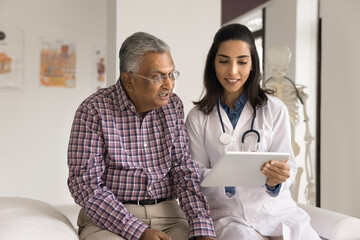 Positive young Indian doctor woman showing electronic content on tablet computer to senior Indian...