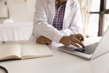 Mature doctor man in white coat typing on laptop at workplace table, holding pen, making medical...