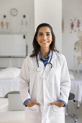 Happy beautiful young Hispanic doctor woman in white uniform coat standing in medical office,...