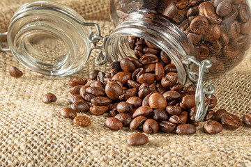 Coffee beans pour out of a glass jar onto a table covered with burlap