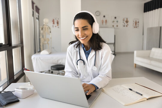 Happy beautiful Latin doctor woman working at laptop in hospital office, typing, smiling, using wireless headphones for video conference call, professional communication, listening to music