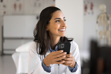 Cheerful young Latin doctor woman holding smartphone in medical office, using healthcare application for communication, giving consultation, support to patients, helping online - obrazy, fototapety, plakaty
